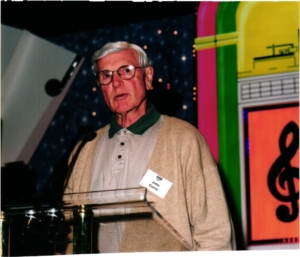 A photo of a man standing at a podium. His nametag says "John Currie"